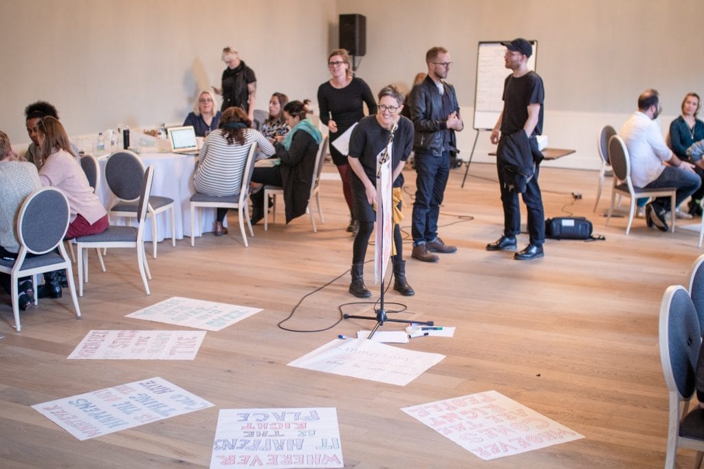 Woman stands in the centre of room. Hand-drawn posters are on the floor around her.