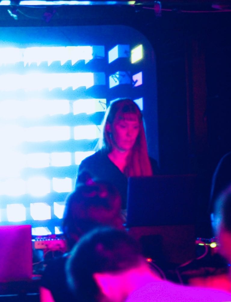 Coral Photoby stands at laptop with light display behind her.