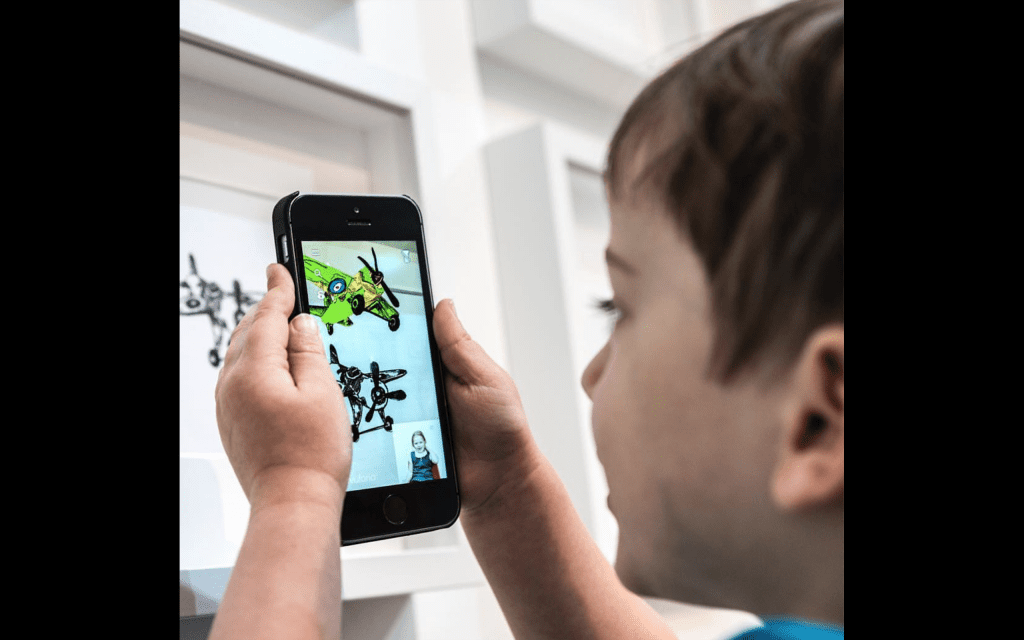 Child holding phone pointed at drawing which turns 3D on screen with someone demonstrating sign language.