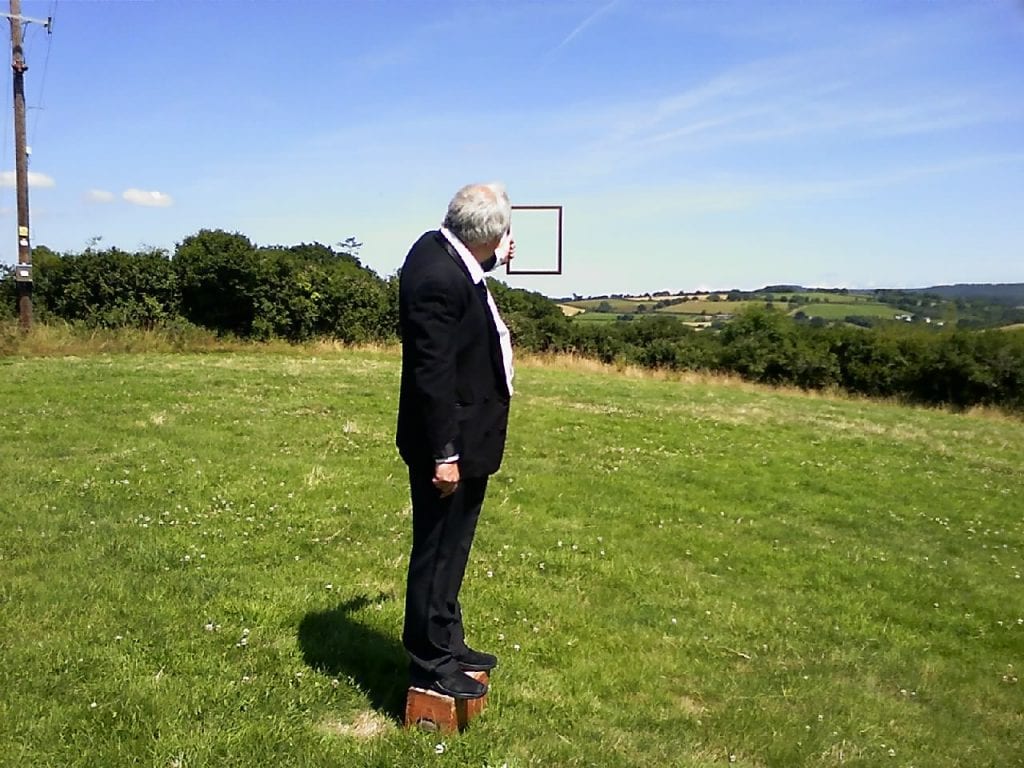 Man in suit stands in field holding up empty picture frame.