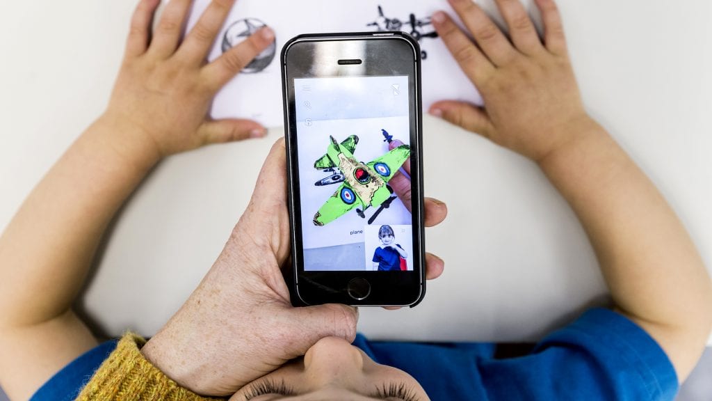 Child's hands on drawing with phone over and screen showing a plane coming out.