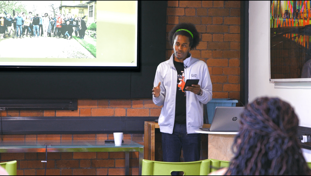 Vincent Baidoo-Lowe stands at front of room speaking to audience with tablet in hand.