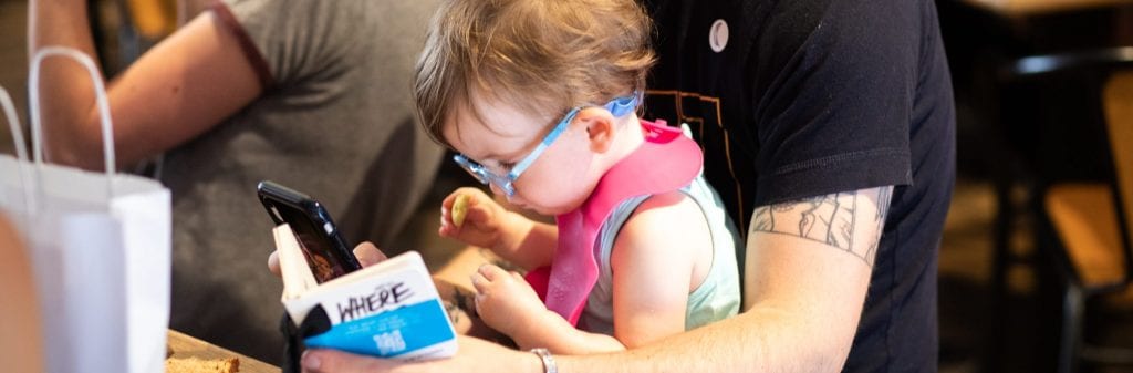 Person holds baby in lap with phone in one hand and small book in the other.