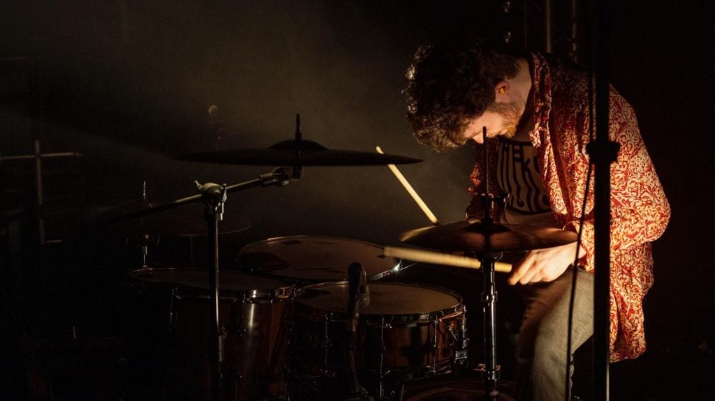 Man in red shirt with head down plays drum kit.