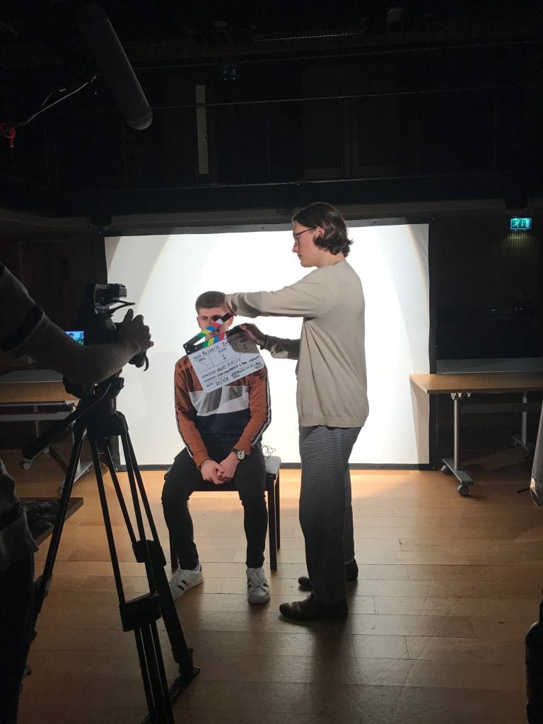 Man sits in front of camera as another man holds clapperboard in front of him.