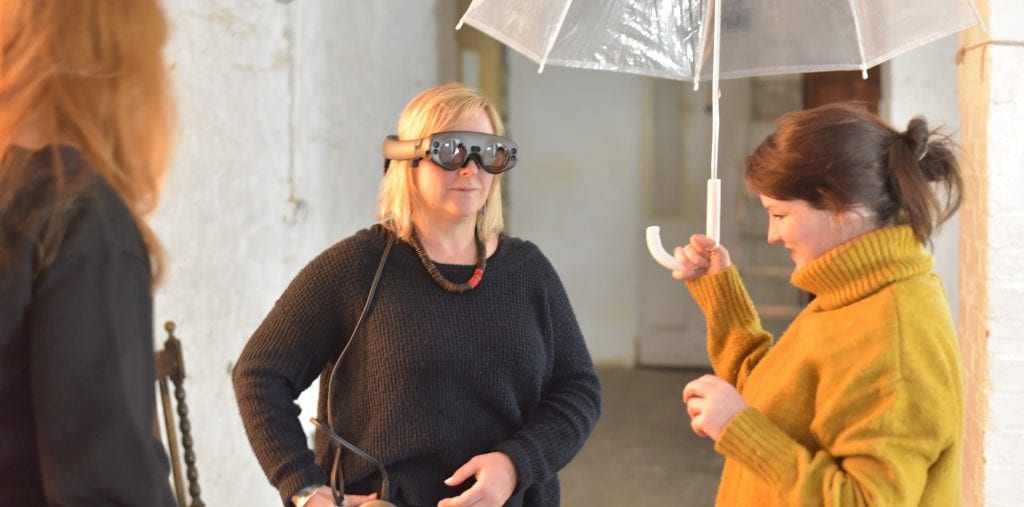 Woman in VR headset stands next to woman holding umbrella.