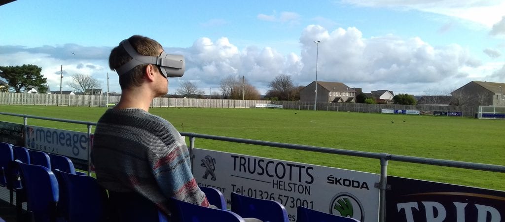 Man in VR headset sits and looks at empty Helston AFC pitch.