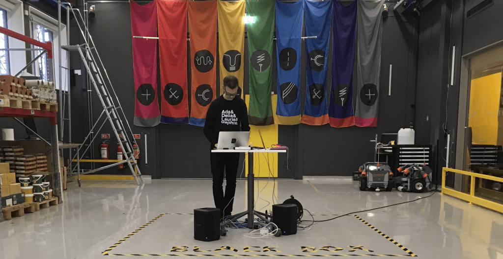 Man stands in room on laptop with colourful flags hanging from wall behind him.