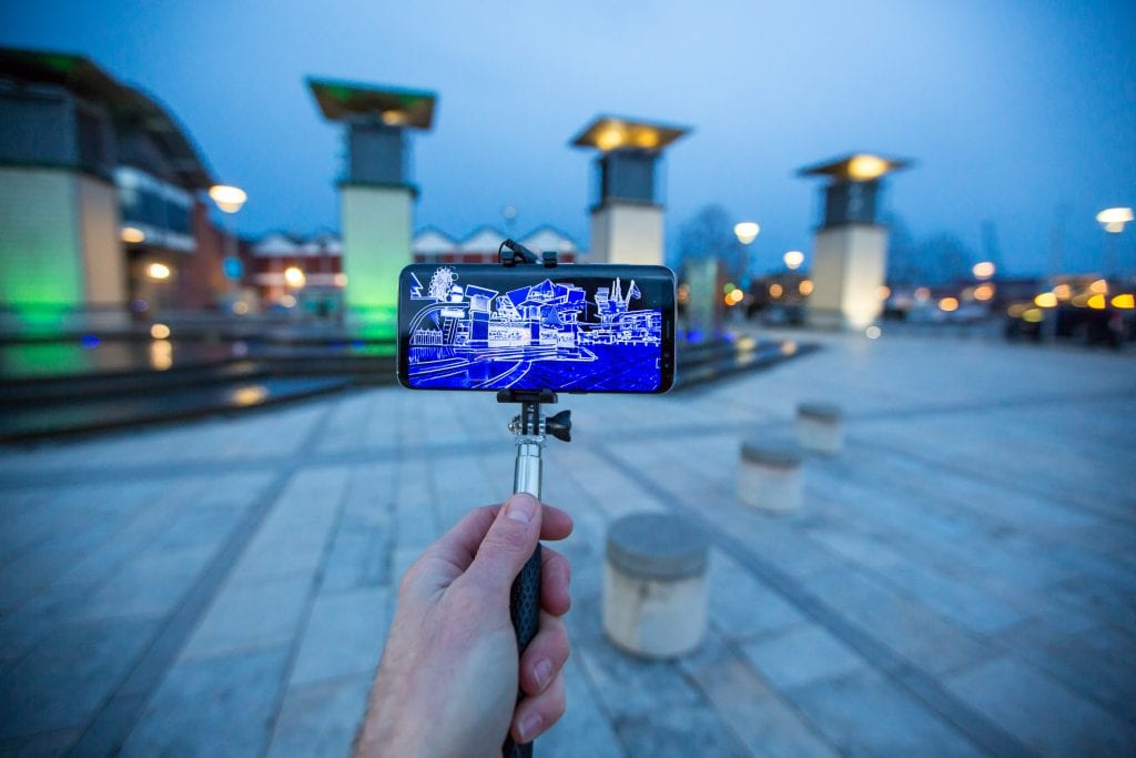 Hand holding selfie stick with phone in facing buildings and showing building plans on the screen.