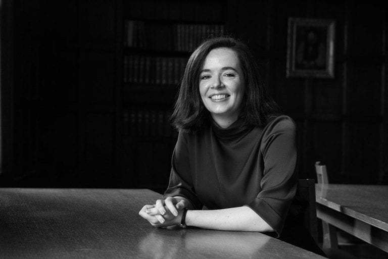 Corrine Stuart sits at a desk smiling, with bookshelf in the background.