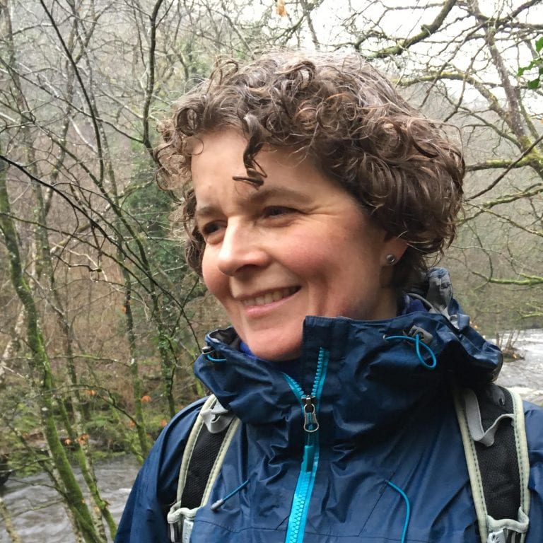 Natasha Nicholson looking away smiling, with woods and a river in the background.