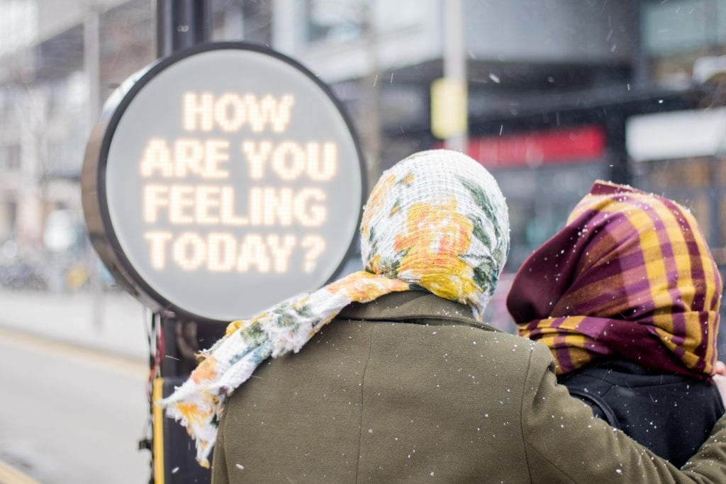 The back of two people's heads wrapped in scarves reading a sign saying "how are you feeling today?"