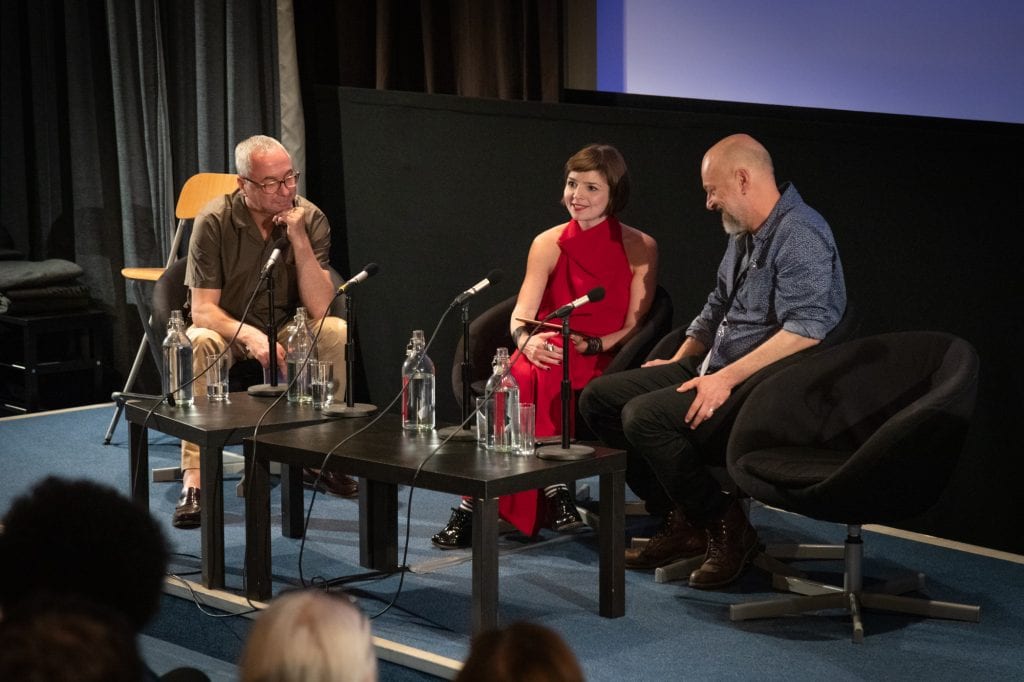 Jane Gauntlett sits between two men during panel discussion listening to audience questions.