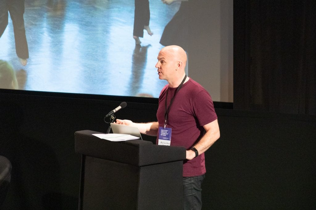 Man in maroon t-shirt stands at lectern in lecture theatre.