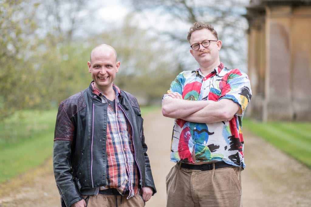 Two men stand outside, one crossing his arms smiling, the other smiling at the camera.