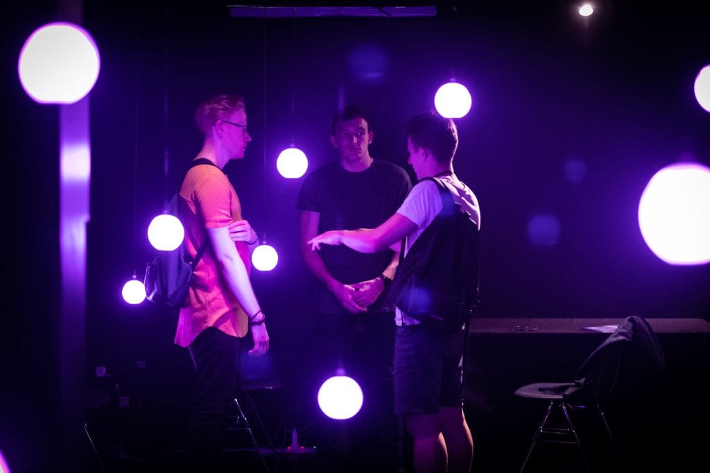 Three people stand in circle talking in dark room with light installation at Squidsoup AudioWAVE showcase