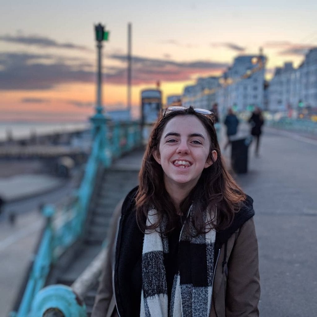 Woman wearing a coat and standing on a seafront with blue railings, smiling at the camera. buildings blurred in the background