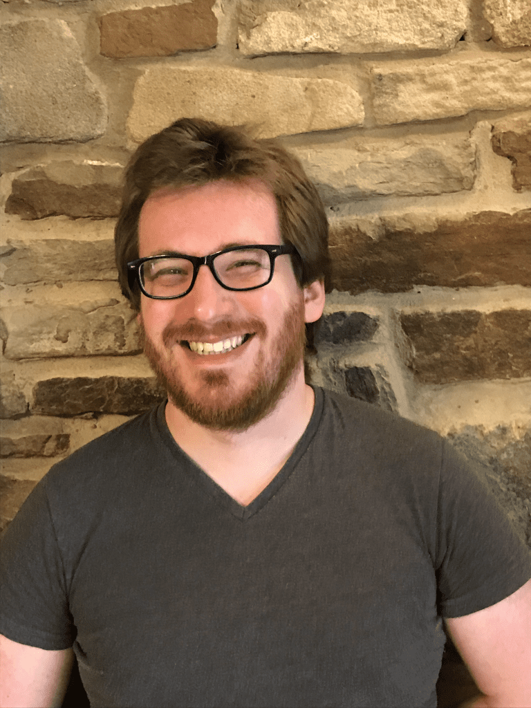 man wearing glasses standing against a wall smiling 