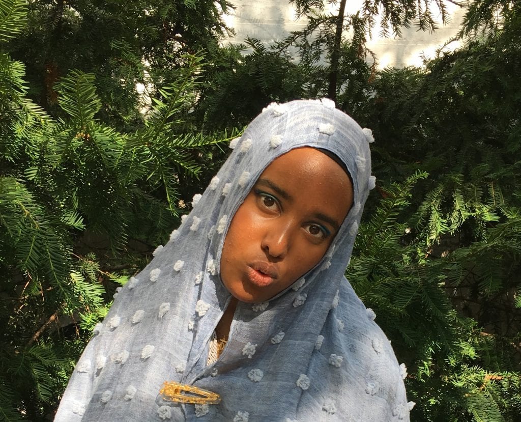 Woman in a blue/grey headscarf, looking directly at the camera, standing against green foliage 
