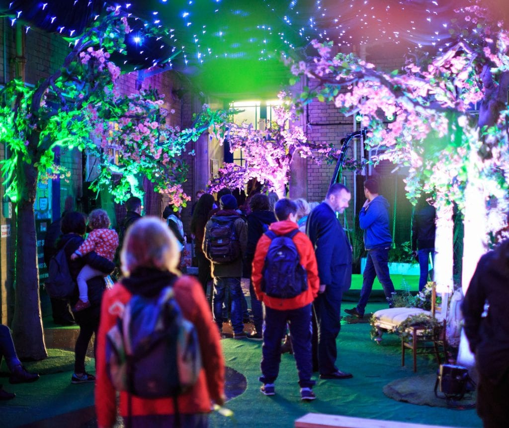 group shot of people walking around an exhibition space, there are trees lit up with coloured lights