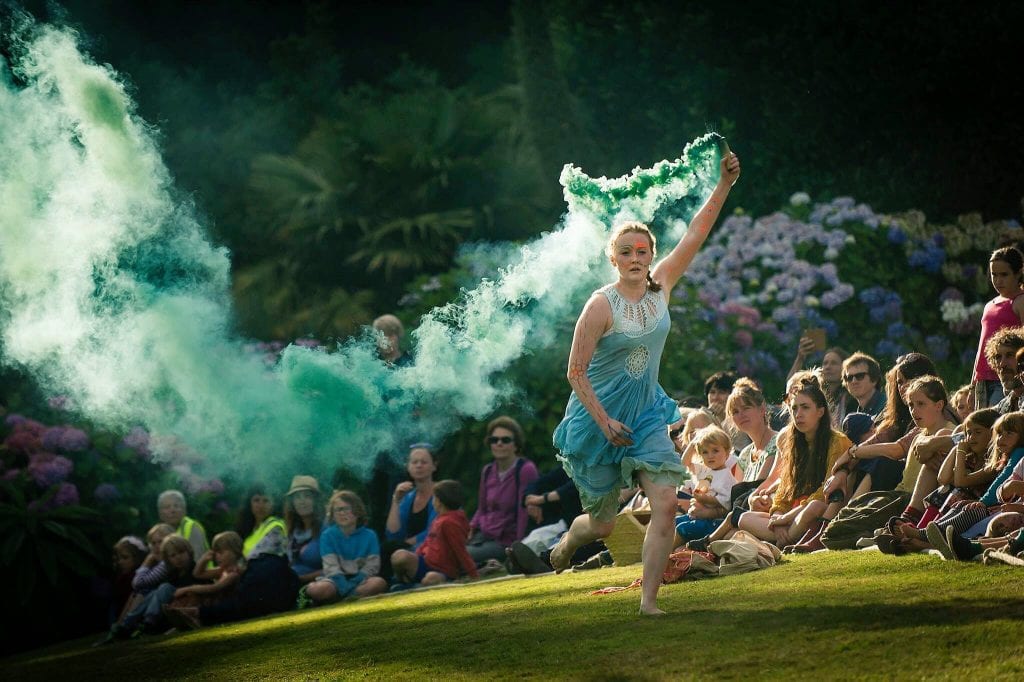 image of a woman running on grass in a light blue dress, she is holding a canister that is releasing teal smoke as a sitting crowd watches 