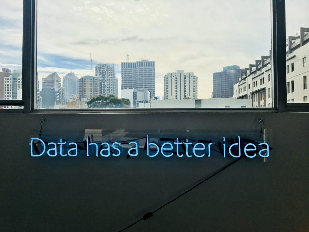 A blue neon sign hanging on the wall written 'data has a better idea'. Above is a widow view of a city landscape.